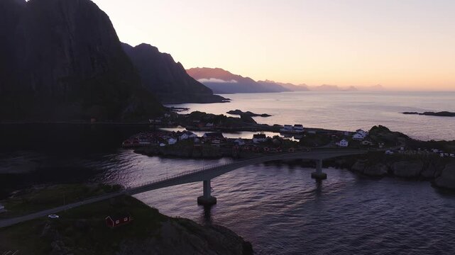 An aerial shot sweeps above a road bridge connecting rocky islands and traditional fishing settlements in Lofoten, Norway during dusk.