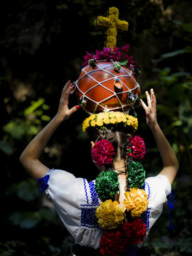 Uruapan, Michoacan, Mexico - 20 April 2025: View of an Aguadora in traditional dress carrying a decorated clay pot on her head, adorned with flowers, a cross, and colorful ribbons.