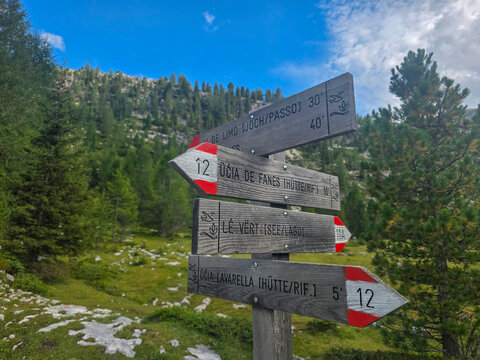 Fanes Dolomites valley Wooden trail signpost pointing directions along a mountain hiking path surrounded by alpine forest, rocky meadow and distant ridgeline under a bright blue sky