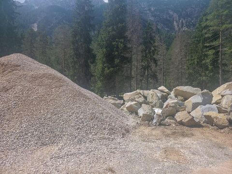 Massive gravel and stone piles in a mountain quarry set against rocky peaks and pine trees, showing texture, scale and raw aggregate material in a rugged natural-construction landscape.