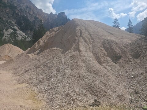 Massive gravel and stone piles in a mountain quarry set against rocky peaks and pine trees, showing texture, scale and raw aggregate material in a rugged natural-construction landscape.