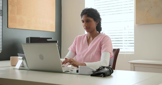 African American woman nurse typing on laptop, pausing to think, laughing in clinic with BP cuff