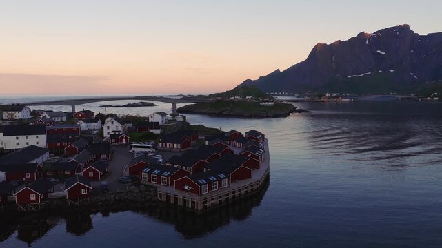 Aerial view captures traditional rorbu cabins along the water in Reine, Norway, with surrounding mountains and bridges during a calm dusk.
