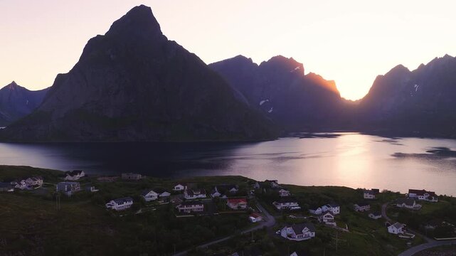 Aerial view gliding over residential houses in Reine, Lofoten, Norway, with steep mountain peaks and fjord water reflecting golden sunset light.