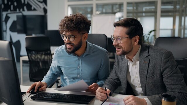 Two multiracial business men analyzing papers review documents charts good result giving high five looking in computer success financial growth multiethnic Caucasian and Arabian businessmen in office