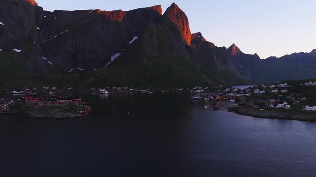 Aerial view over the fjord water towards traditional rorbuer cabins and steep mountain peaks glowing in early morning light in Reine, Lofoten, Norway.
