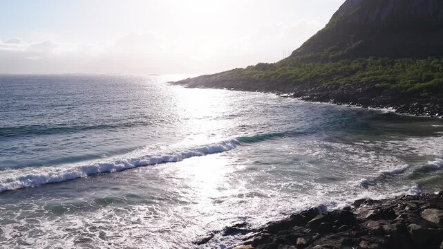 Aerial view flying over ocean surf as waves break along the rocky coastline and mountain slope of Lofoten, Norway under bright sunlight.