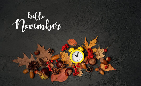 Hello November greeting card. alarm clock and fall nature decorations - leaves, nuts, cones, acorns on dark background. November month calendar concept. Daylight saving time end. flat lay