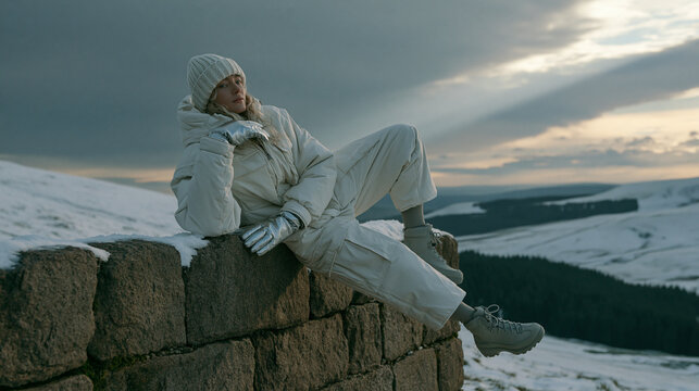 Person in wei&szlig;er Winterjacke sitzt auf Steinmauer vor verschneiter Berglandschaft, ruhige Wintersaison-Stimmung