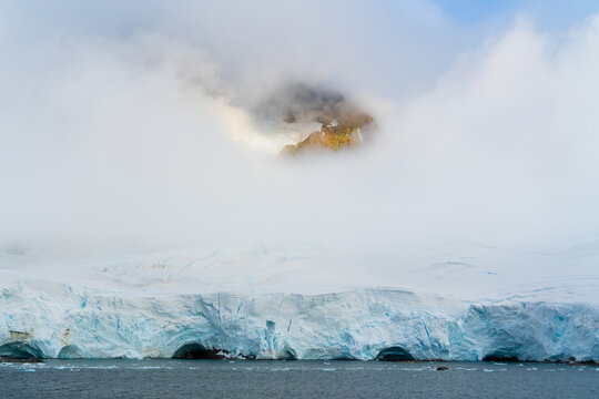 Antarctica: a grandiose landscape characterized by an imposing blue ice cliff, surmounted by a thick layer of snow and low clouds that reveal a rocky peak.
