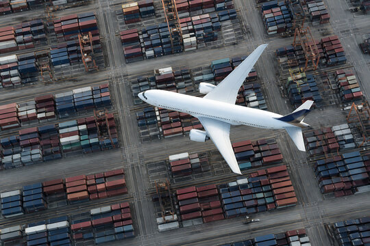 A white airplane is flying over a large, dense container yard in an industrial setting.