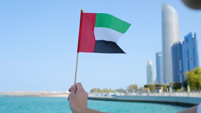 Female hand holding small UAE flag against Abu Dhabi skyline. Symbol of national pride and modernity with iconic skyscrapers in the background showcasing urban elegance and progress