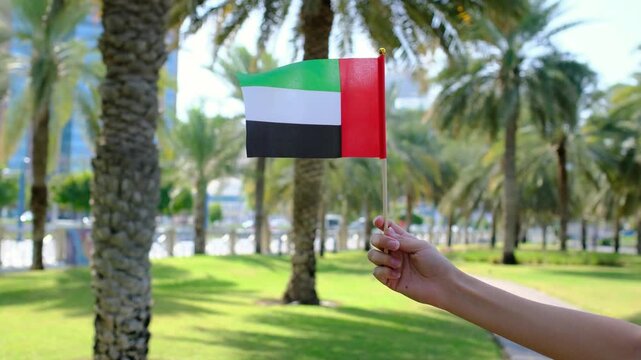 Female hand holding small UAE flag against Corniche Walking Trail in Abu Dhabi. Blend of nature, patriotism, and serenity, with lush greenery , views symbolizing harmony and progress.