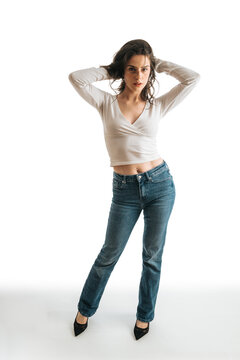 Young woman wearing jeans and a white top poses confidently with hands behind her head. Casual studio portrait of a model standing against a white background in a relaxed pose.