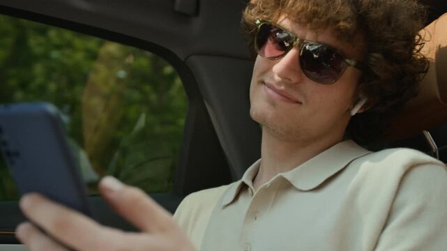 Portrait of Caucasian young adult man wearing sunglasses and earbuds, sitting in car, lowering sunglasses to look at camera while using smartphone. Perfect for mobile app advertisements