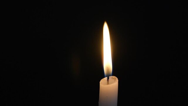 Close up of a lit white candle. Small flame moving on a black background.