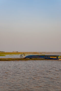 Kampong Phluk, Siem Reap Province, Cambodia - 05 February 2020: View of a blue boat beached on the shore of Tonle Sap lake with a bicycle nearby and a stilt house in the distance.