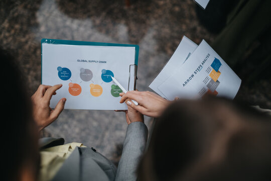 Business professionals review a global supply chain chart on a clipboard outdoors. Colleagues discuss documents and arrow steps analysis while pointing at diagrams during an outside meeting.