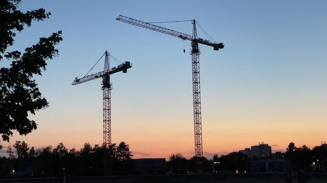 two tall tower cranes rise over construction site at twilight, their lattice masts and long jibs outlined against clear sky, city skyline fading into soft evening