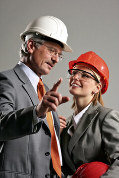 Bauleiter im grauen Anzug mit wei&szlig;em Schutzhelm erkl&auml;rt Ingenieurin mit orangem Helm etwas, Business-Foto f&uuml;r Baubranche und F&uuml;hrungsthemen.