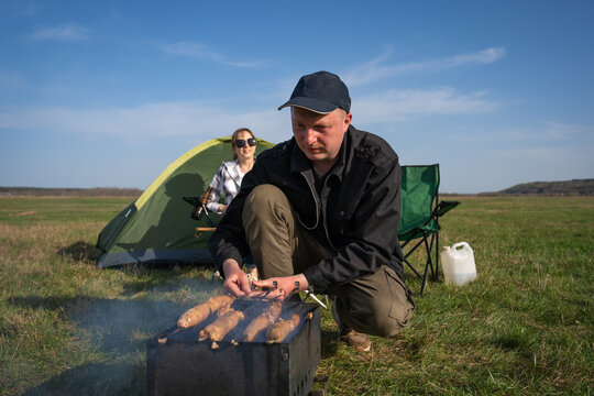 Man squatting at a portable grill cooking sausages over coals and smoke while woman relaxes in a camping chair beside a tent in a grassy field on a sunny day