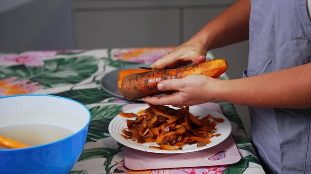 A person is peeling a carrot on a kitchen table with a patterned tablecloth, viewed from a top-down perspective. The scene is set in a well-lit, clean kitchen with a neutral mood, ideal.
