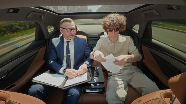 Middle aged Caucasian businessman and young adult Caucasian partner arguing over documents in backseat of moving car, young man tearing paper. Video ideal for corporate conflict or legal dispute