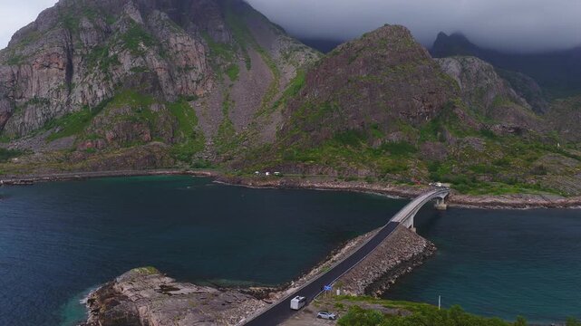 An aerial perspective showcases a highway bridge connecting rocky shores across sea waters against towering steep mountains in Lofoten, Norway.
