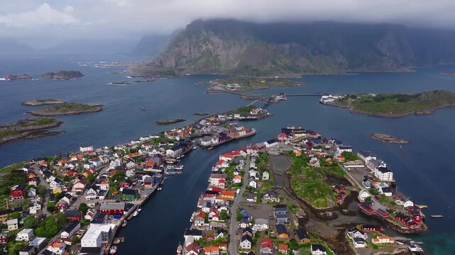 Aerial video moving over the coastal town of Henningsvaer in Lofoten, Norway, showing traditional houses, sea channels, bridges, and distant mountains.