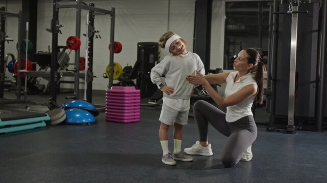 Caucasian female trainer kneeling next to Caucasian child in gym, guiding him through stretching exercises. Perfect for promoting family wellness programs or childrens fitness classes