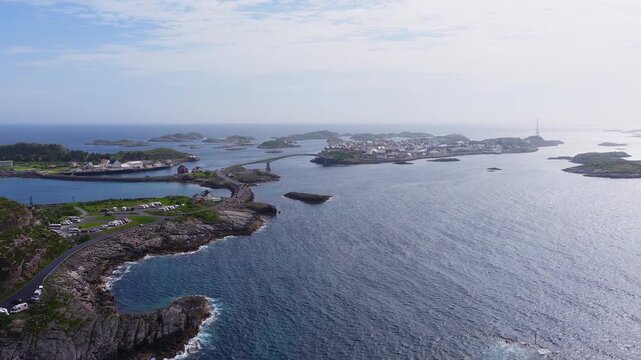 An aerial view reveals the road and bridge connecting the rocky islands to the fishing village of Henningsvaer in Lofoten, Norway under a bright summer sky.