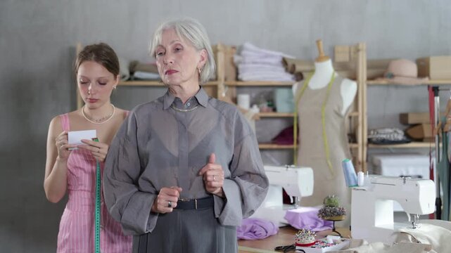 Attentive young female dressmaker measuring mature woman using tape in sewing workshop