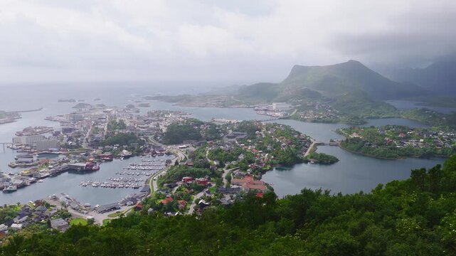 An elevated aerial view shows the coastal town of Svolvaer in Lofoten, Norway, surrounded by fjords, islands, mountain ridges, and harbor marinas.