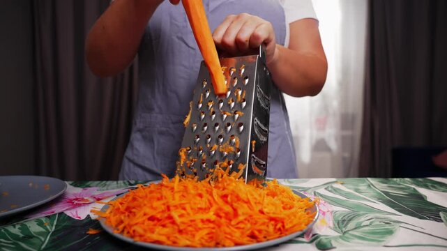 A person is grating carrots using a grater in an indoor setting with curtains and a tablecloth. The scene is captured from a close-up, slightly elevated angle, with a neutral mood.