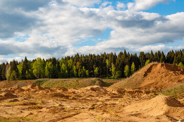 Expansive view of a textured sand quarry featuring piles of loose soil under a dramatic blue sky with fluffy white clouds, bordering a dense coniferous and deciduous forest backdrop © PhotoChur