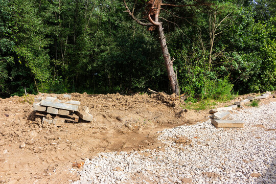 Rough road construction site with piles of brown dirt, gravel roadbed, and stacked concrete curbstones at the edge of a forest