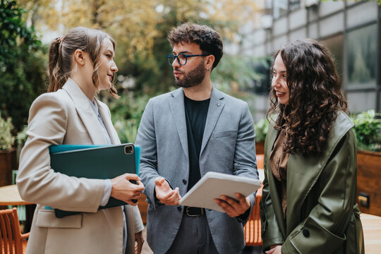 Three colleagues discuss work while reviewing a tablet and folder during an outdoor business meeting. They stand in a courtyard talking and sharing documents.