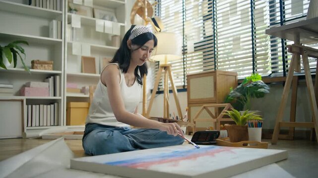 Young Asian woman artist painting on canvas easel with paintbrush in art studio, representing creative professional women designer, fine arts student lifestyle and freelance illustrator