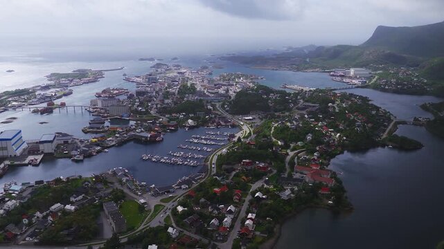 Aerial view of the coastal town Svolvaer in Lofoten, Norway, showing the harbor, boats, residential buildings, roads, bridges, and sea under an overcast sky.