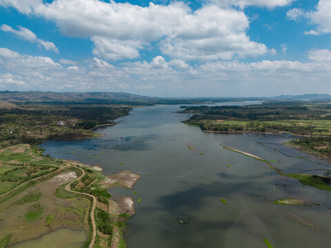 Aerial view of the Sengguruh Reservoir surrounded by lush green fields, agricultural land, and rolling hills under a bright blue sky in Malang, East Java, Indonesia.