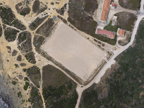 Aerial view of a rectangular sandy sports arena surrounded by dirt paths and coastal vegetation near a building in Portugal.