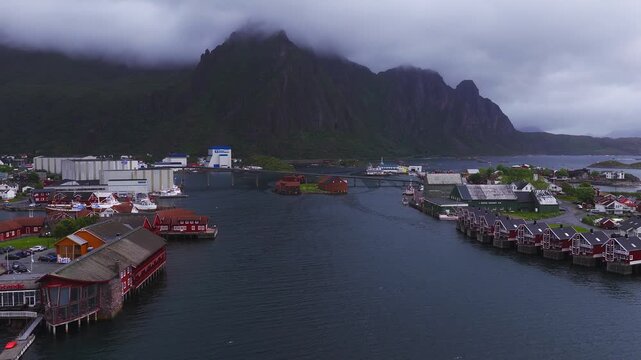 An aerial perspective flying above the coastal waters of Svolvaer harbour in Lofoten, Norway, showing traditional waterfront houses and mountains.