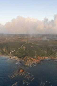 Aerial view of rugged coastal cliffs meeting the Atlantic Ocean with rolling hills and soft clouds in the sky in Portugal.