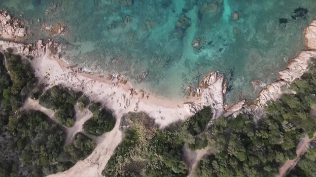  Aerial video starting from a top-down straight view of a wild sandy beach and clear turquoise water ripples, flying forward over the sea and slowly tilting up to reveal a scenic panorama of a rocky r