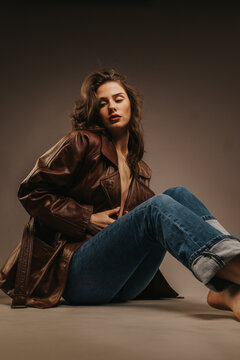 A young woman wearing a brown leather jacket and rolled jeans poses barefoot against a neutral studio backdrop. Studio fashion portrait of the model in casual outerwear and a relaxed seated pose.