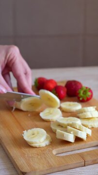 Female hands slicing banana with knife on wooden board. Close up of woman hands cut banana on cutting board, preparing healthy meal.