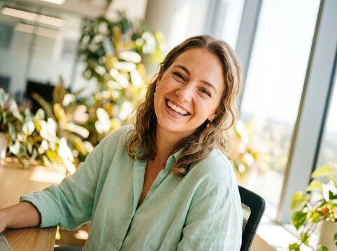Smiling woman sitting in bright office surrounded by green plants, showing warmth and confidence during relaxed professional moment
