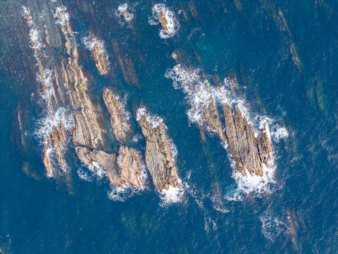 Aerial view of jagged rock formations rising from the deep blue ocean with white sea foam and waves crashing against the rugged stone surfaces in Portugal.
