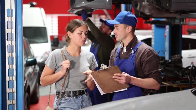 Positive young auto-mechanic in blue overalls communicating with woman client of auto-shop about service fees indicated on sheets