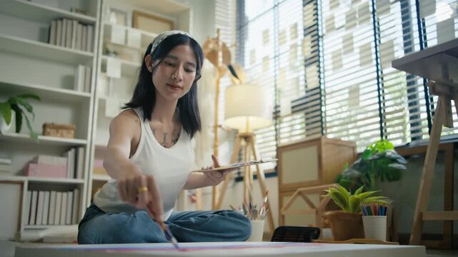 Young Asian woman artist painting on canvas easel with paintbrush in art studio, representing creative professional women designer, fine arts student lifestyle and freelance illustrator